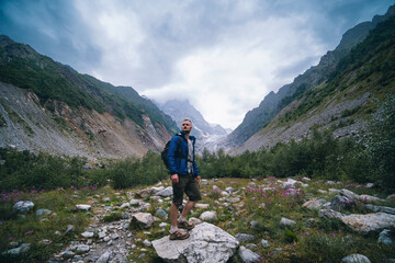 Fototapeta premium Traveler man with a backpack standing on a stone, On the way, walk the track. A valley among majestic high mountains.. Back view. The Chalaadi Glacier, Georgia, Caucasus. Blue jacket.