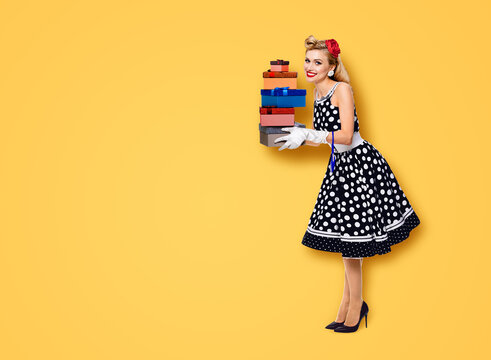 Big sales discounts, rebates offers ad concept - full body image of happy smiling beautiful woman in pin up black dress with polka dot, white gloves, hold gift boxes, isolated orange yellow background