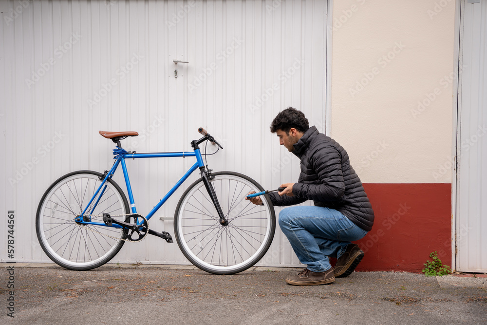 Wall mural Man wearing jeans and black coat fixing a blue bike in front of garage