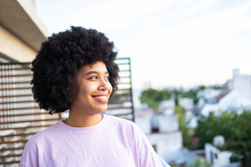 Happy Latin girl with afro hairstyle looking away . She is wearing pink shirt on the apartment balcony and enjoying the view