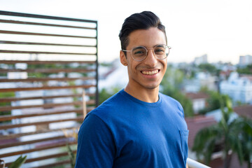Smiling Hispanic man with glasses wearing blue shirt smiling on the apartment balcony with view of the city. 