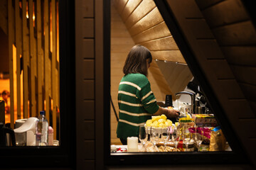 Woman prepares dinner in the kitchen, a view through the window of a country house at night.