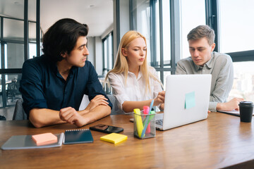 Concentrated young male and female startup business colleagues having discussion sitting at desk looking at laptop screen in light office room by window. Concept of corporate work meeting .