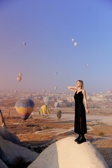 Traveler woman in black dress background landscape Cappadocia with hot air balloons sun light. Concept trip Turkey travel
