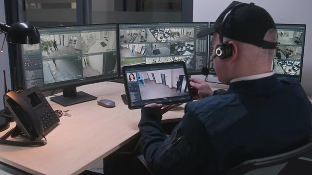 Security officer in headset controls and monitors CCTV cameras in coworking office, uses digital tablet and computers with playback on screens. High tech software showing surveillance cameras footage.