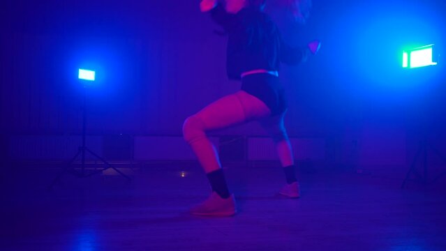Blonde girl with ponytail wearing black hoody and shorts dancing twerk in misty dance studio, blue flashlight on background. Provocative dancer shaking buttocks. Concept of choreography