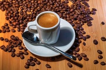 A cup of rich espresso on a saucer with a spoon, surrounded by roasted coffee beans on a wooden table, perfect for a coffee shop promotion. 