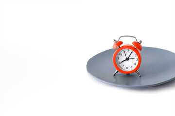A red alarm clock stands on a plate on a white background. The concept of diet and proper nutrition on a schedule. Bright red alarm clock and gray plate. Free space for text and ads