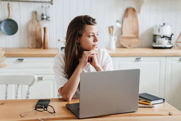 Pensive hispanic young adult woman sits at table with laptop at kitchen exhausted of hard work. Sad Italian girl feels fatigue looks aside thinks about financial troubles. Overloaded female, failure. - obrazy, fototapety, plakaty