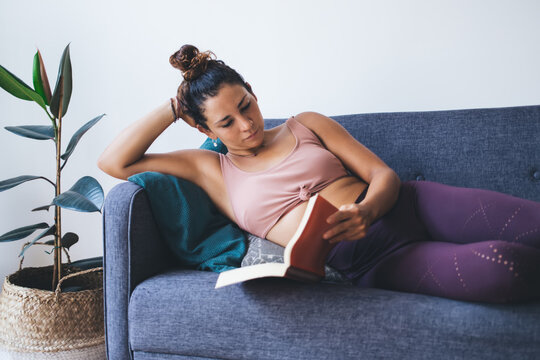 Interested young woman holding literature book and relaxing at home, Caucasian hipster girl reading bestseller with thriller plot resting on comfortable sofa in apartment with stylish flat interior