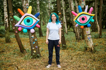 Weird young adult woman with dyed turquoise hair in white T shirt near big eyes art installation in...