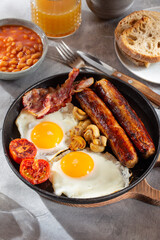 Full English breakfast with fried eggs, sausages, beans, bacon, grilled tomatoes, mushrooms, toasts and cup of coffee on gray background.