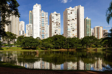 Fototapeta premium Paisagem do lago no Parque Flamboyant com alguns prédios atrás das árvores e imagem dos prédios refletidas na água.