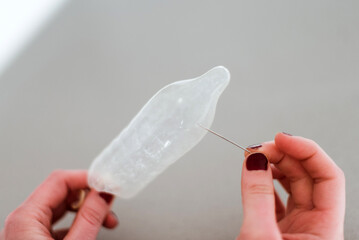 Una mano femenina pinchando con una aguja un preservativo de látex. Hacer un agujero en los preservativos para provocar el embarazo. Fallo del método anticonceptivo.  - obrazy, fototapety, plakaty