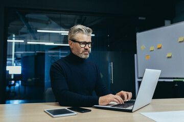 Serious mature gray-haired businessman working inside modern office, man thoughtfully typing on laptop at workplace.