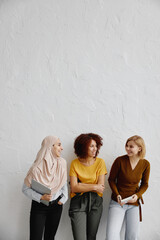 Three elegant smiling young women with different ethnicity standing over white wall background.