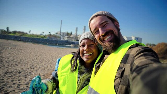 A environmental activist or volunteer multiracial couple picking up trash og the beatch, and taking a selfie portrait smiling looking at camera showing a recycle plastic bag. High quality 4k footage - Powered by Adobe
