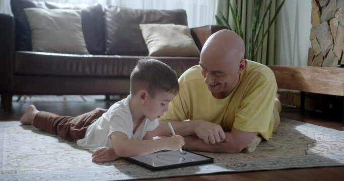 Happy family father drawing on graphic tablet helping cute son enjoying talking game lying on warm floor at home, dad and little boy having fun in living room at leisure. 4K