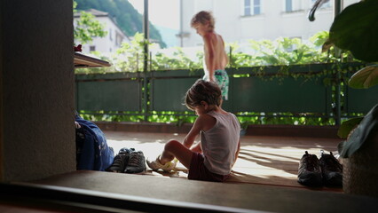 Child putting shoe by doorstep home entrance. Kid sitting on floor puts tennis shoes