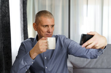 A man with tea makes a selfie photo with a mobile phone in the apartment.