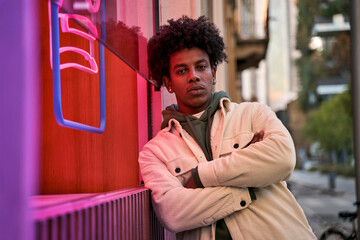 Confident cool African American hipster guy standing at city street near neon light sign. Rebel gen z male model looking at camera outside advertising young generation culture life entertainment. - Powered by Adobe