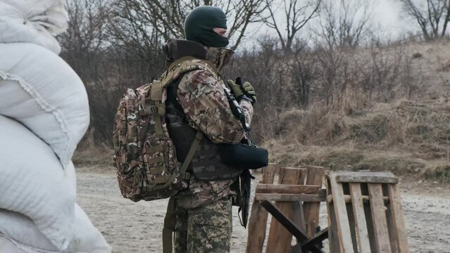 Soldier with weapon in military uniform stands in front of the barricades from sandbags and anti-tank hedgehog barriers. Military man on the roadblock. Combatant in full ammunition. Concept of war