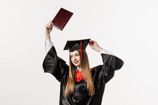 Happy girl student in black graduation gown and cap raises masters degree diploma above head on white background. Graduate girl is graduating college and celebrating academic achievement.