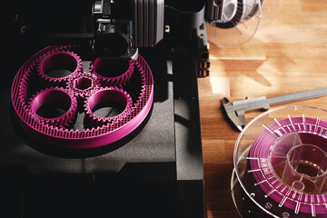 Black FDM-printer makes planetary gear machine part with visible infill from pink filament. Top view on printer, tools and filament roll on wooden desk area. Modern production concept. Selective focus - obrazy, fototapety, plakaty