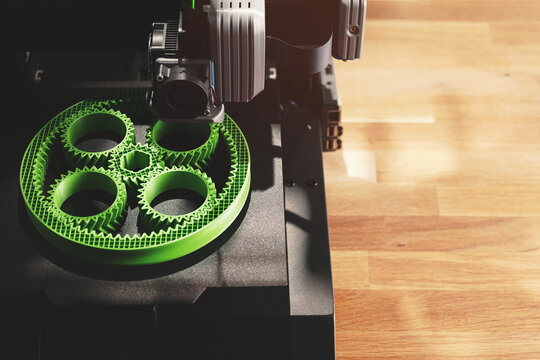 Black FDM-printer makes planetary gear machine part with visible infill from green filament in bright light. Top view on printer on wooden desk area. Modern production concept. Selective focus