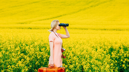 Stylish girl in pink dress with vintage camera and travel suitcase watching in binoculars in...