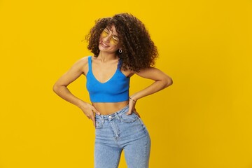 Woman with curly afro hair in a blue T-shirt on a yellow background dancing flying hair with sunglasses yellow, hand signs, look into the camera, smile with teeth and happiness, copy space