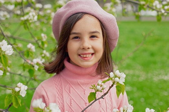 Portrait of happy little girl stands in a blooming white apple tree in the park.
