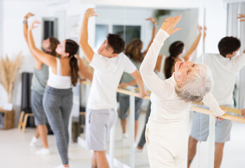Gracile old-aged woman engaging in ballet at ballet barre in training hall during workout session