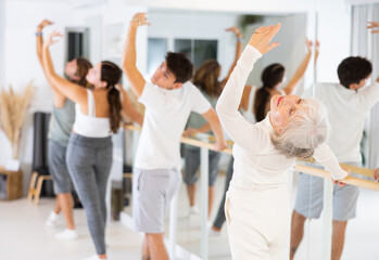Fototapeta premium Gracile old-aged woman engaging in ballet at ballet barre in training hall during workout session