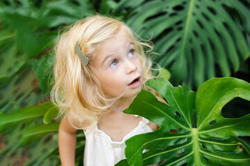 Cute adorable caucasian pretty blonde two year old girl, toddler, kid among big green monstera leaves. Natural beauty,perfect skin, health,cosmetics concept.Close up shot.