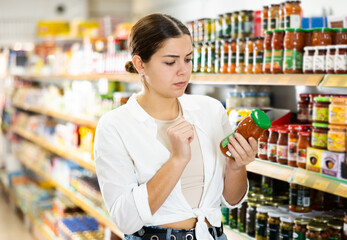 Interested thoughtful young female shopper choosing food in supermarket, holding glass jars with...