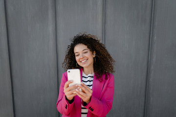 pretty curly smiling woman walking in city street in stylish jacket, using smartphone