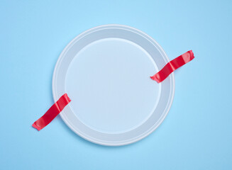 Empty white plastic plate attached with red adhesive tape on a blue background, a concept of...