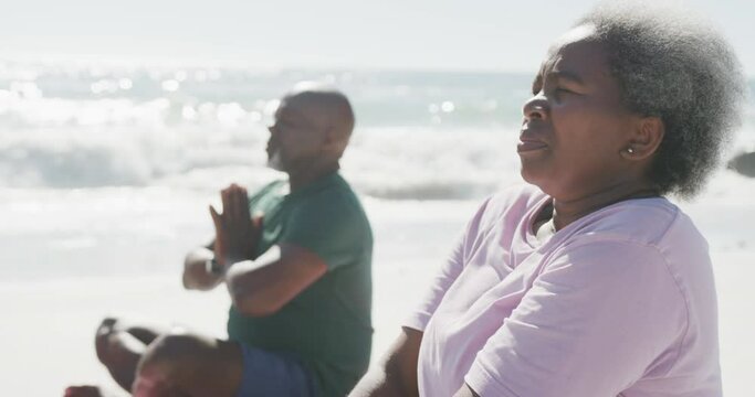 Happy senior african american couple doing yoga and meditating at beach, in slow motion