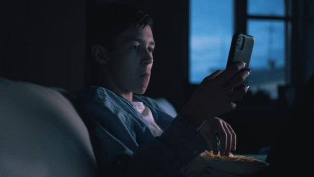 Happy Teenage Boy Using Phone and Eating Chips at Home - Nighttime Leisure and Socializing