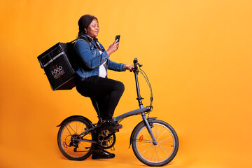 African american womandelivery standing on bike in studio with yellow background while checking client adreess on phone before start delivery takeaway meal. Pizzeria courier carrying take out backpack