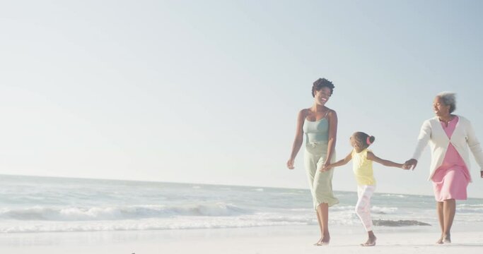 Happy african american grandmother, mother and daughter walking at beach, in slow motion, copy space