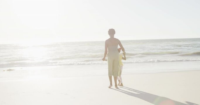 Happy african american mother and daughter walking at beach, in slow motion