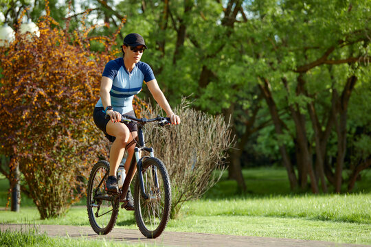Smiling middle aged woman in cap with glasses cycling in summer park, cycling cardio training, city walks with bike, weight control