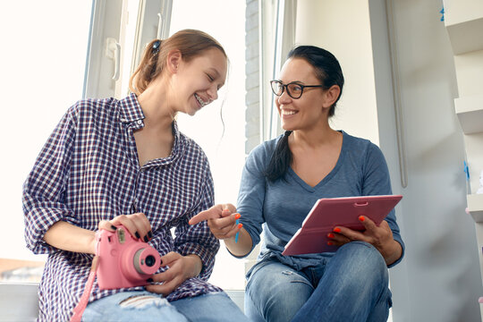 Happy young mother and teenage daughter have fun sitting by the horse in the kitchen. Smiling mother and daughter holding camera and albos with family photos in hands, enjoying family life together at