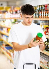 Focused thoughtful young guy choosing food in supermarket, holding glass jars with canned goods and reading labels