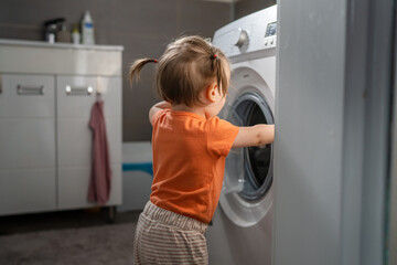 Fototapeta na wymiar One girl small caucasian toddler child daughter standing at the washing machine in the toilet opening or closing the door examine and learn early development and growing up mischief concept copy space
