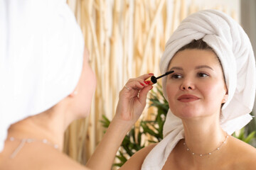 An attractive young woman paints her eyelashes with mascara in front of the bathroom mirror. 