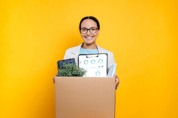 Positive mixed race young woman with glasses got a new job, holding cardboard box, standing on...