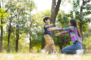 Fototapeta premium Asian little cute boy running to hug his mother at the park, mother and sun relaxation activity in a park or garden in weekend. Woman and a boy playing on a beautiful sunlight and bokeh background.